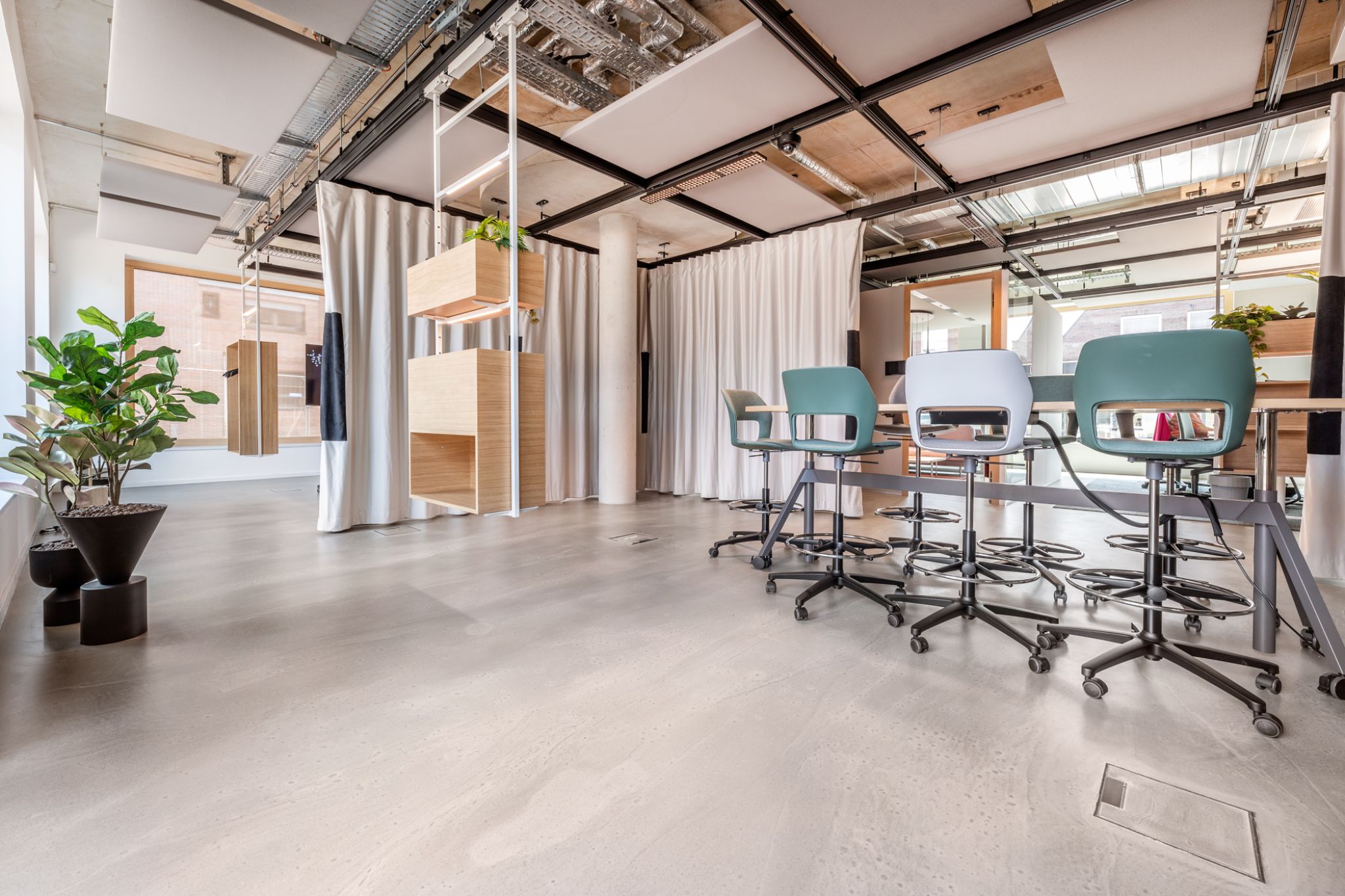 Modern office space with wooden and metal furnishings, gray swivel chairs, potted plants, and ceiling ductwork. Natural light enters through large windows, illuminating the area.