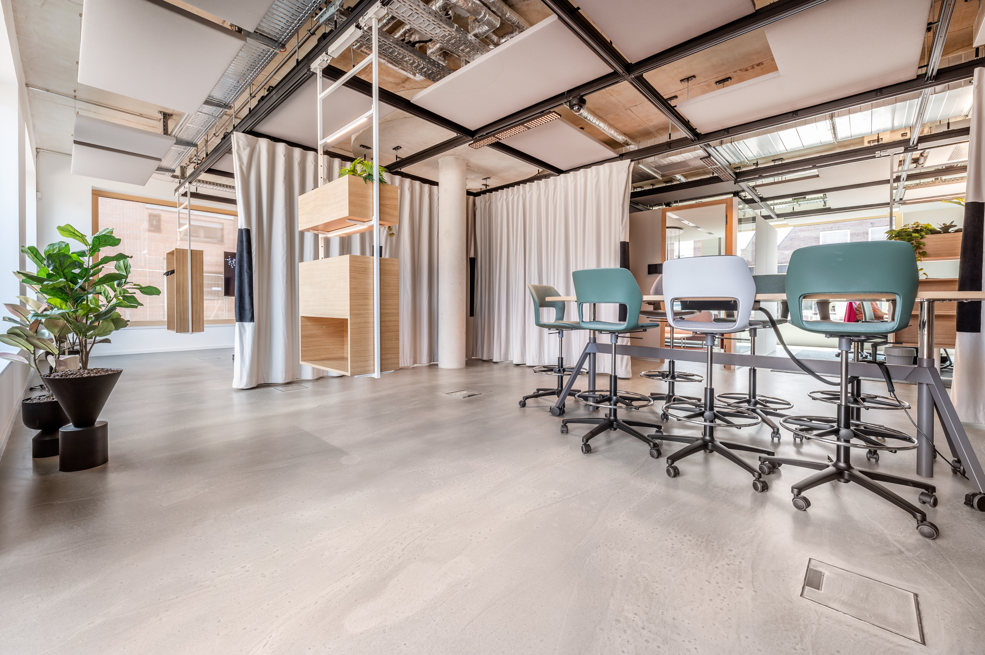 Modern office space with wooden and metal furnishings, gray swivel chairs, potted plants, and ceiling ductwork. Natural light enters through large windows, illuminating the area.