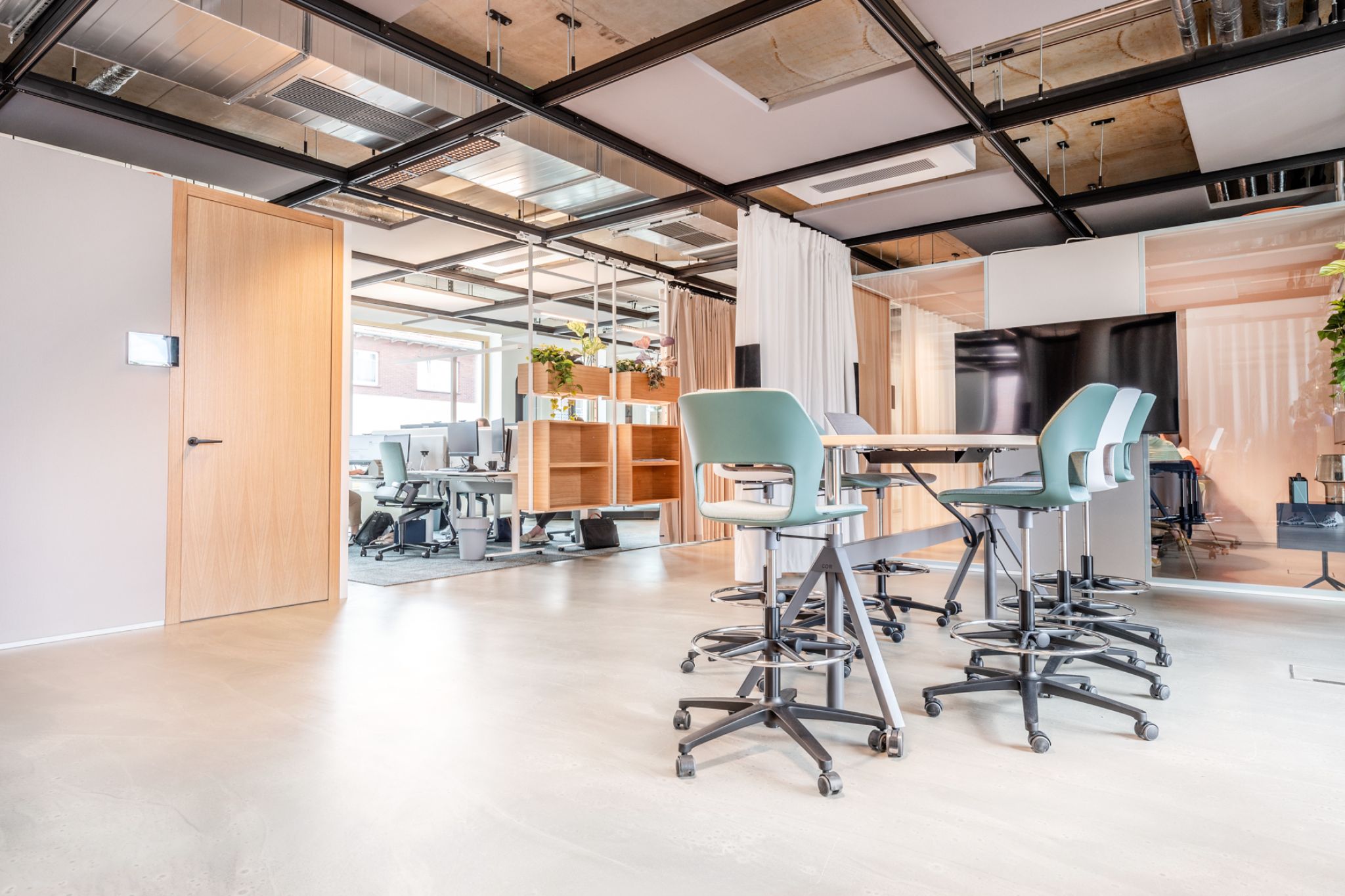 A modern office space with high chairs around a table, open workstations, wooden door, and natural light from windows.