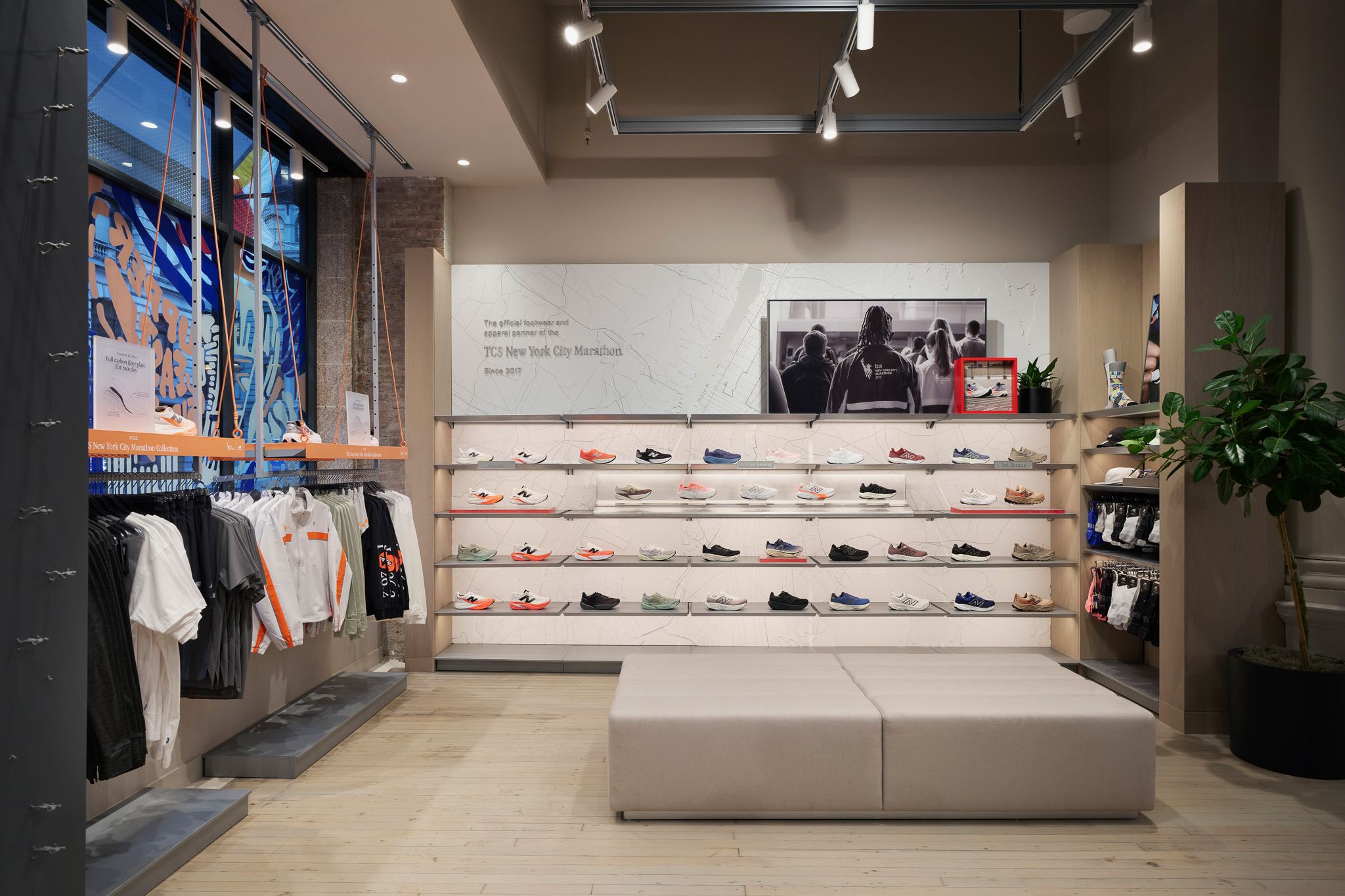 Interior of a shoe store with wall shelves displaying sports shoes, a central bench, and clothing on the left wall.