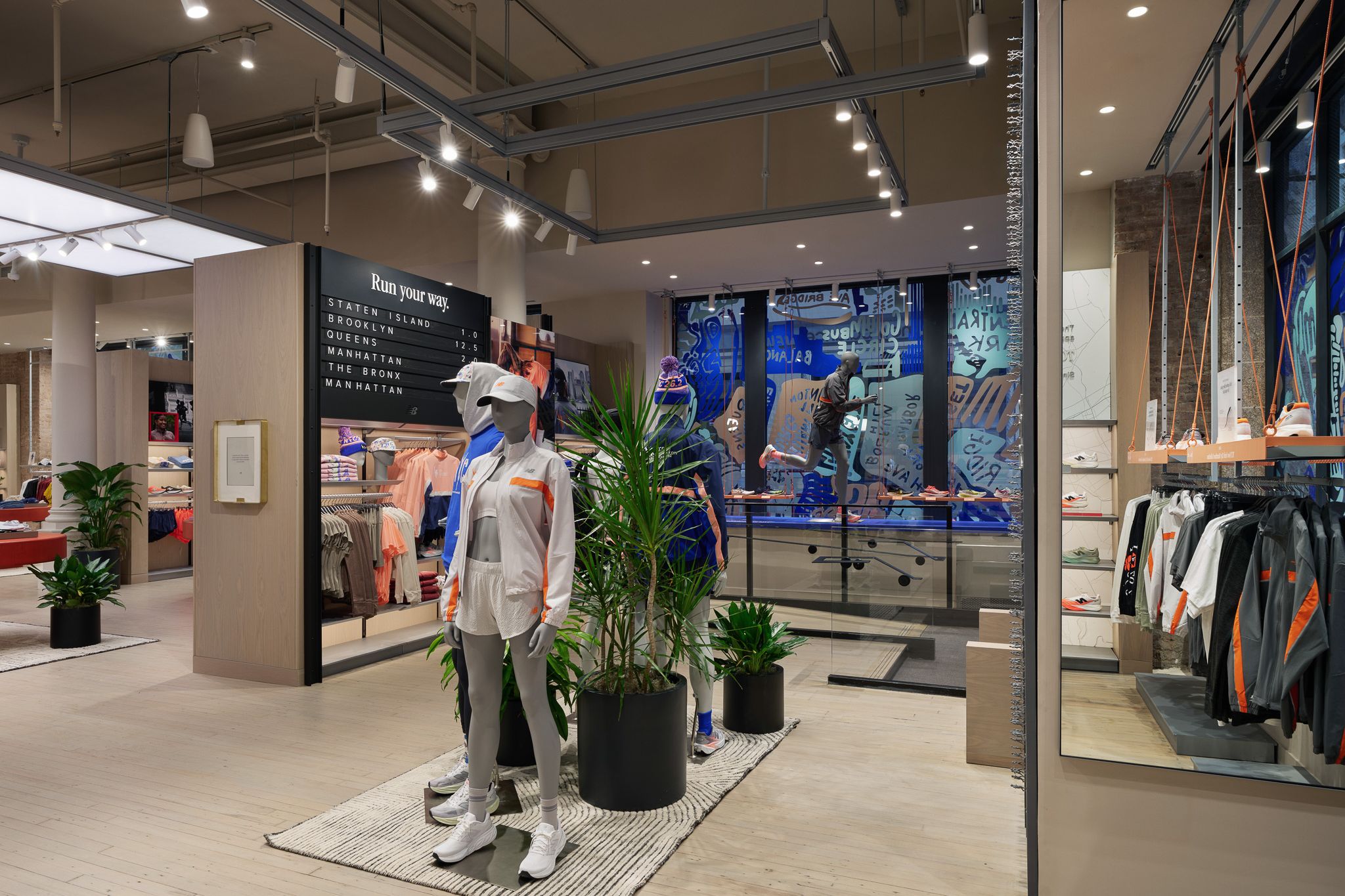 Two mannequins dressed in sporty clothes next to green plants in a clothing store with light wood floors and ceiling lights.